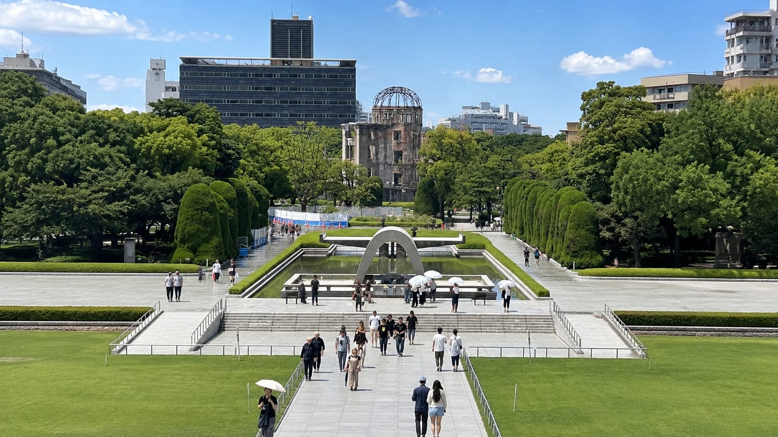 Photograph of Hiroshima Peace Memorial Park on a clear summer day, looking north along the central axis: the arched Cenotaph frames the Pond of Peace with the skeletal Atomic Bomb Dome aligned in the distance, and visitors walk the paved approach under parasols.