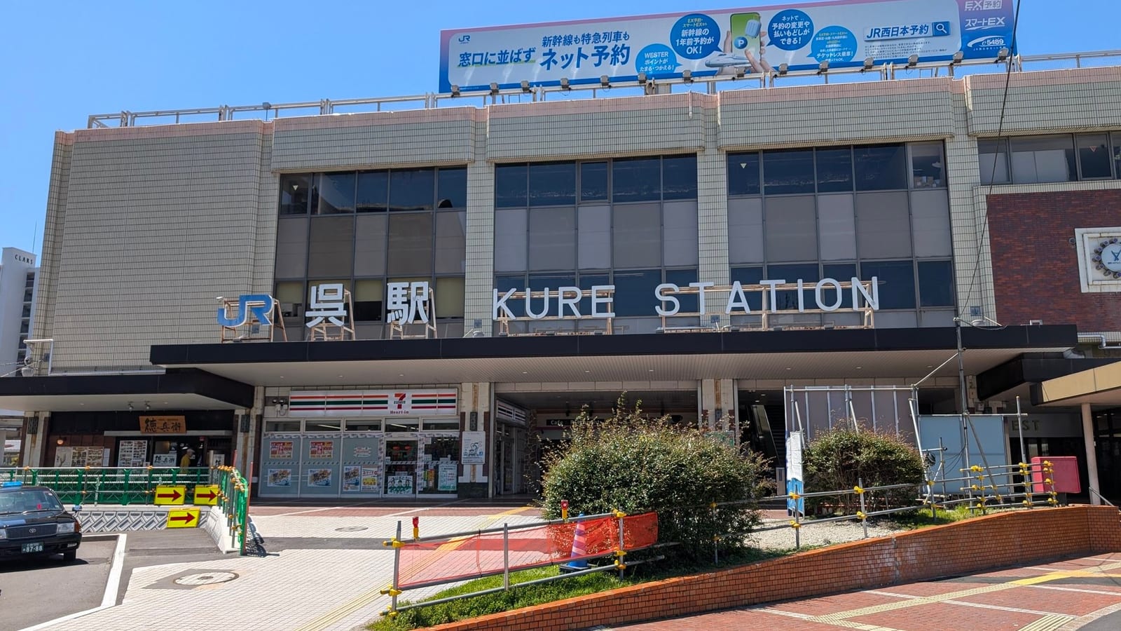The front of JR Kure Station on a clear day, with the KURE STATION sign above the entrance and a 7-Eleven at street level.