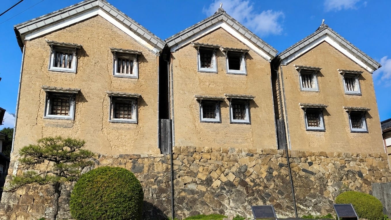 The three Mitsukura storehouses of the former Sawahara residence in Naganoki-cho: earthen-walled kura standing in a row on a rough stone base, seen from the street.