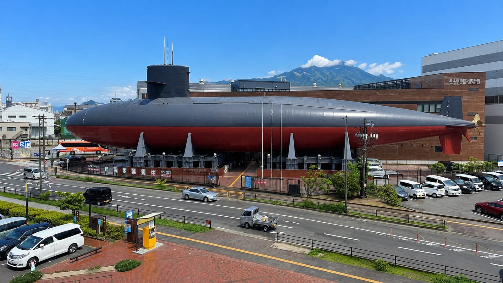 The submarine JS Akishio mounted beside the JMSDF Kure Museum, seen from the elevated pedestrian deck on the walk from Kure Station, her black upper hull and red underside raised on steel cradles above street level.
