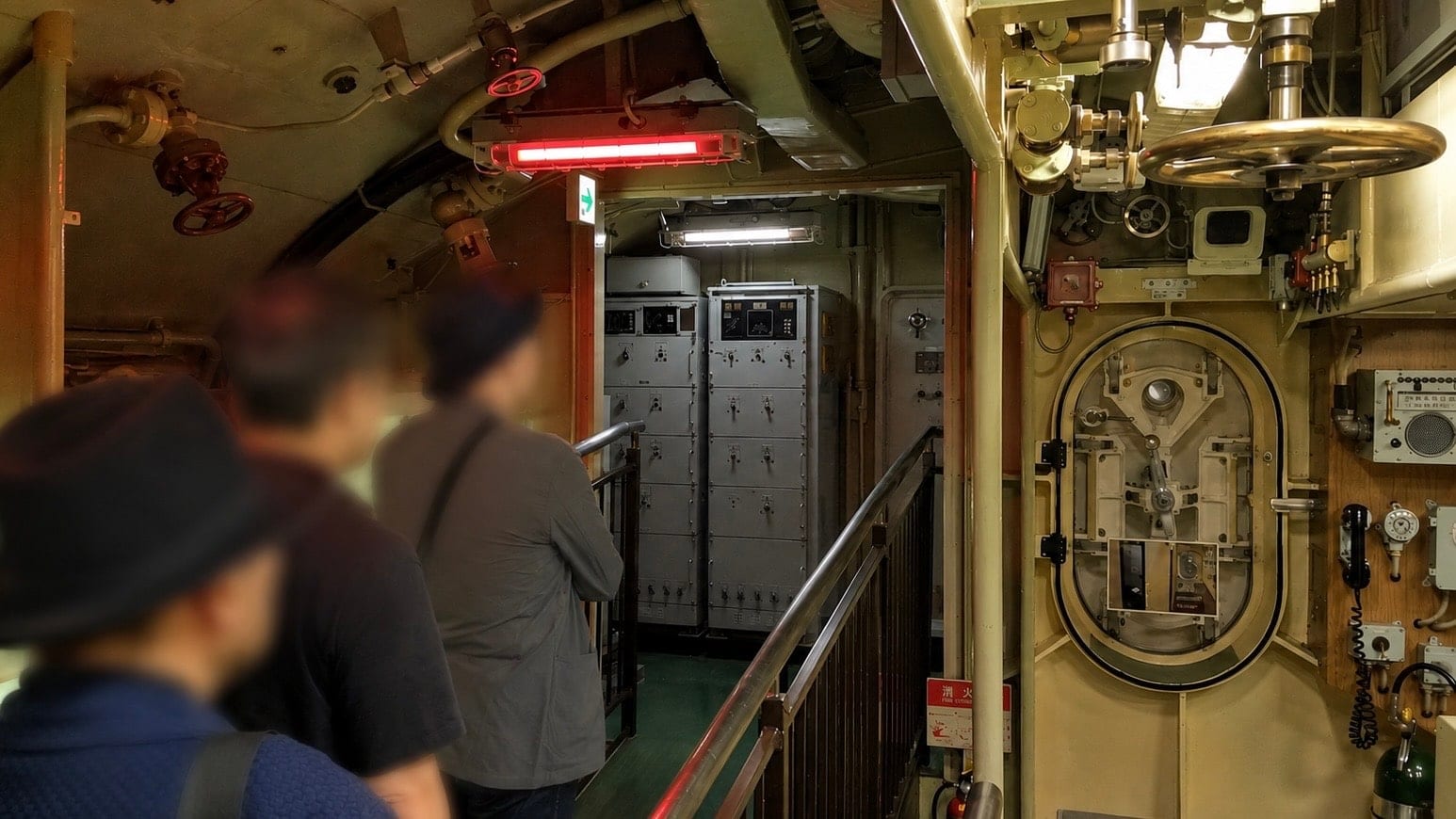 Inside the JS Akishio: visitors file single file through a narrow passage past equipment cabinets, with a watertight door and valve wheels on the right and pipework overhead.