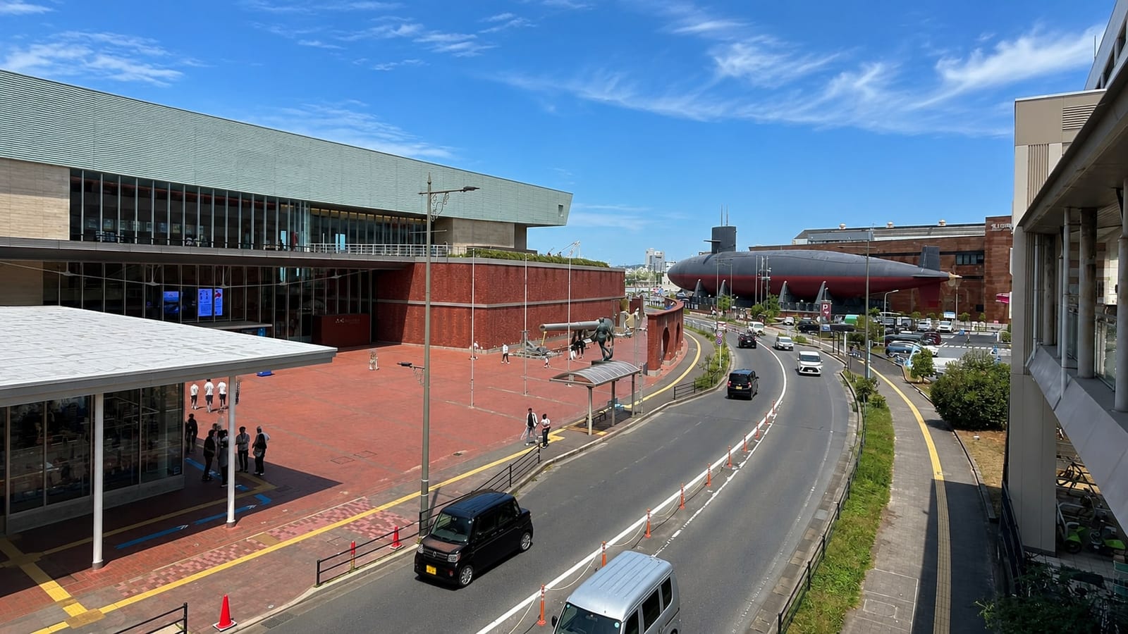 Both museums in one frame from the elevated walkway: the Yamato Museum and its brick plaza on the left, with the battleship Mutsu's gun barrel lying beside the road, and the submarine Akishio across the street on the right.