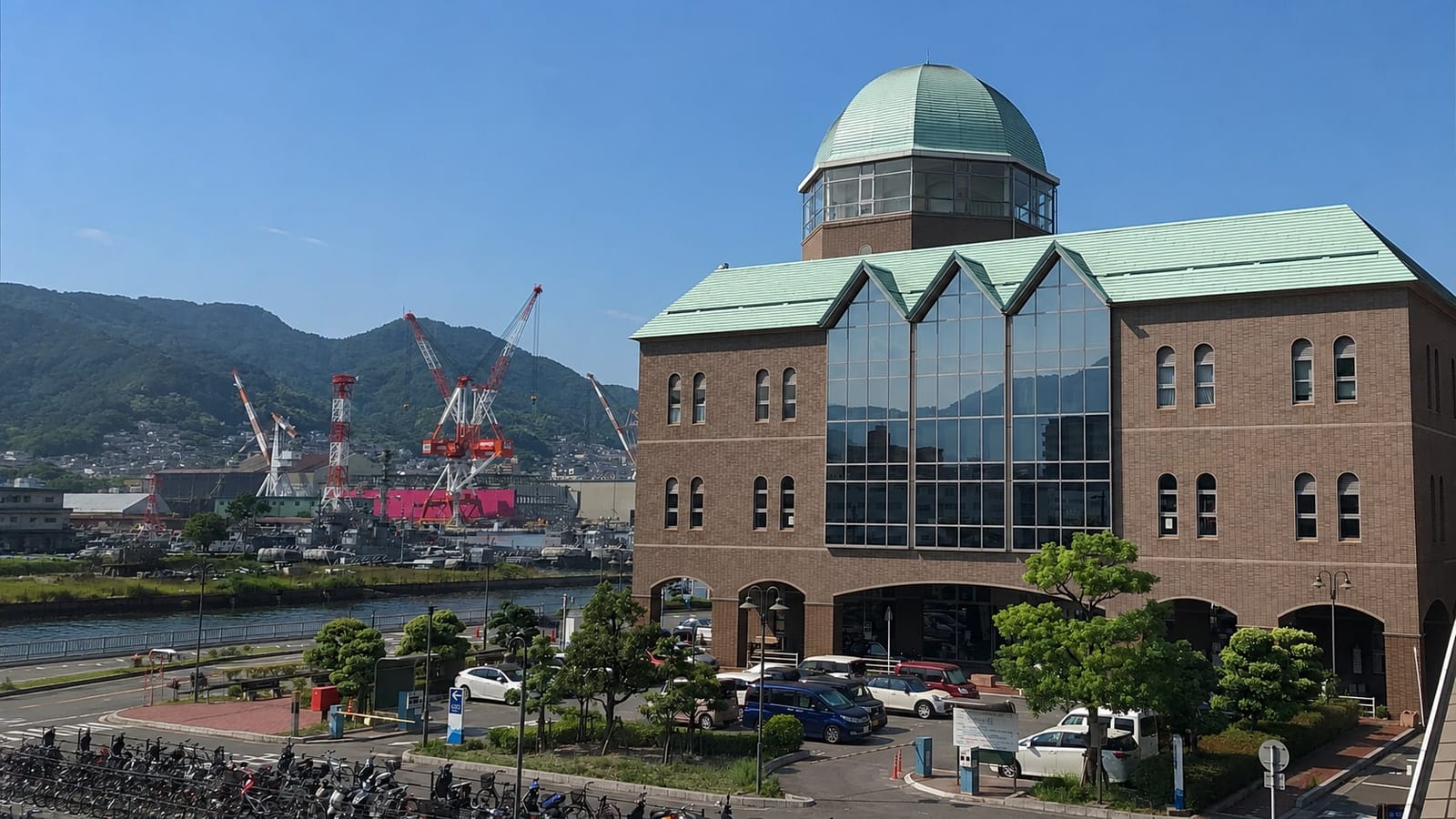 The Kure Chuo Sanbashi Terminal — a brick building with a glass atrium and a green dome — with the shipyard cranes and grey JMSDF vessels visible behind it.