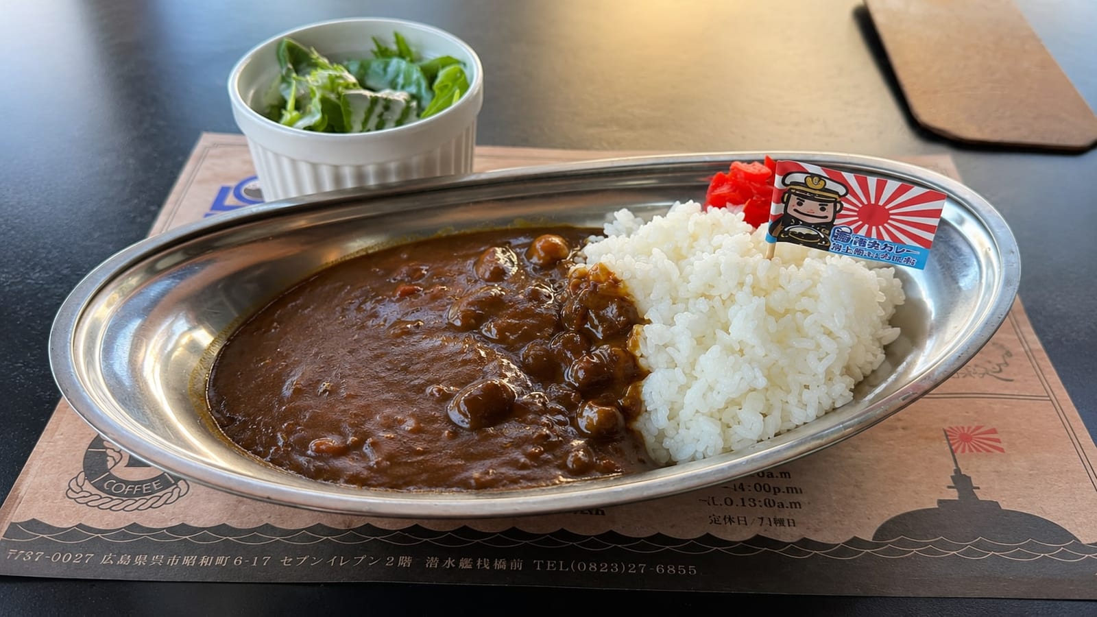 The JS Kuroshio recipe curry at Minato Machi Coffee Ten — soft beef curry and rice on an oval steel plate with a small side salad, on a paper mat printed with the shop's address above the 7-Eleven, in front of the submarine pier.