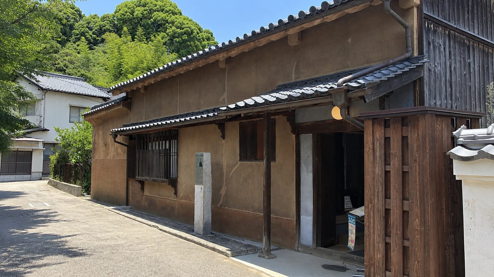 The former Kaneko Family Residence in Mitarai, an Edo-period merchant house with pale earthen plaster walls and a tiled roof, its wooden nagaya-mon gate at right and a stone interpretive marker set at the lane in front.