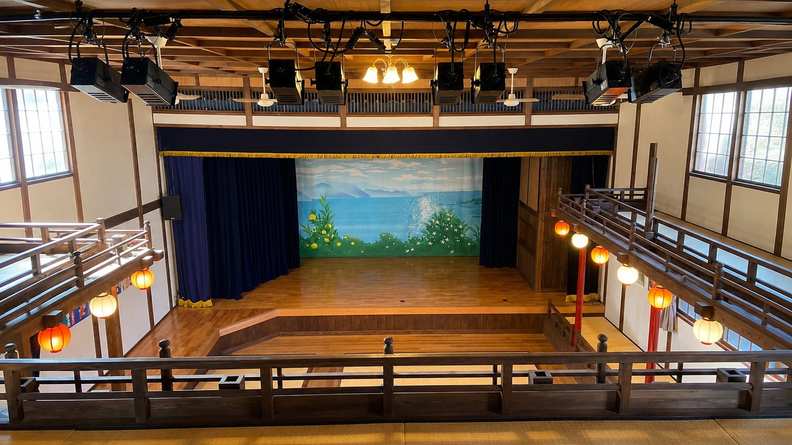 The interior of Otome-za, Mitarai's small 1937 wooden theatre, seen from the upper gallery toward the stage, with tatami seating below, red and white paper lanterns along the balcony rails, and a painted Seto Inland Sea backdrop on the stage.