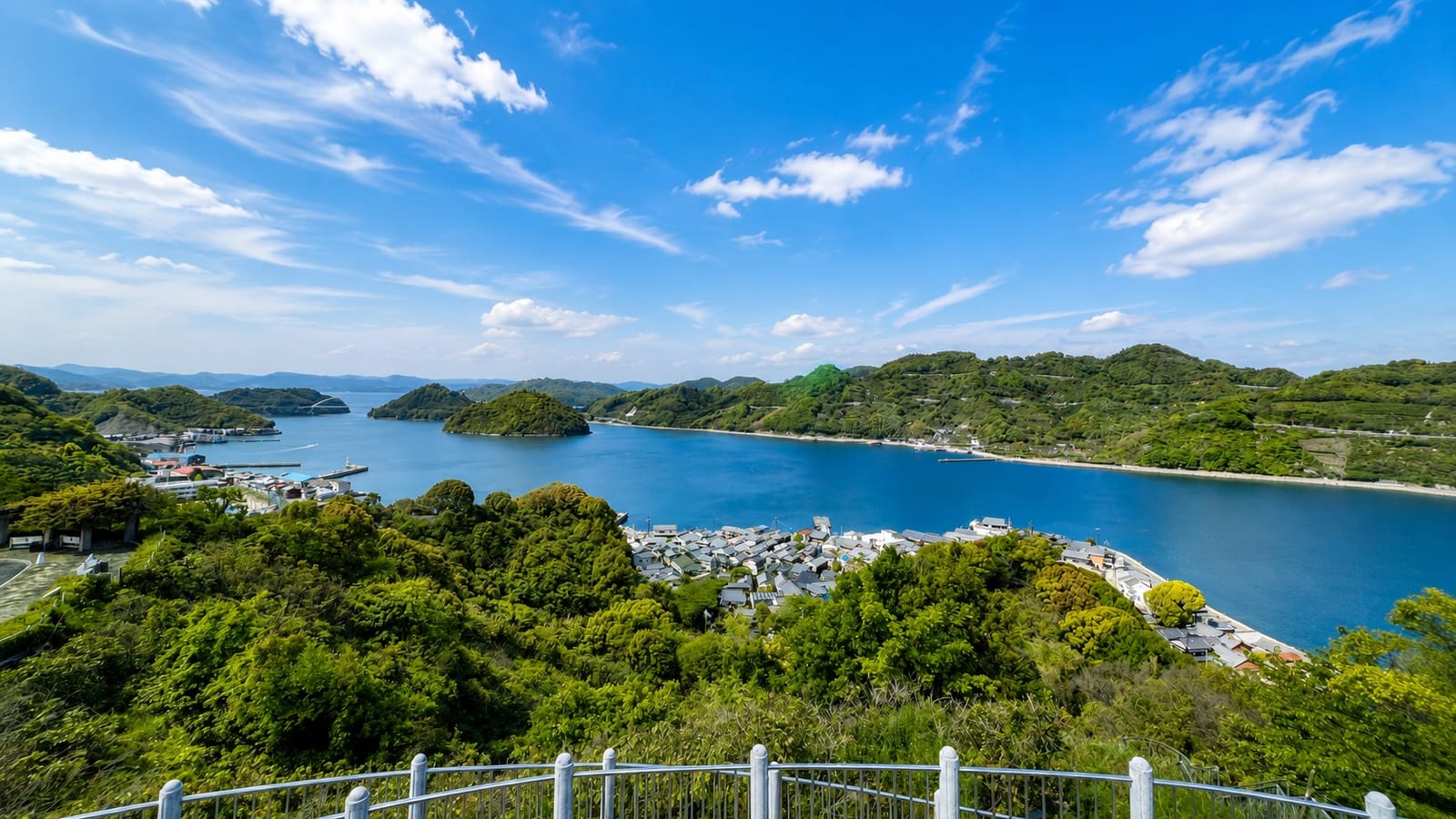 Mitarai seen from the hillside lookout above the town, the tiled roofs of the old port in the foreground and the narrow blue channel between Osaki Shimojima and the neighbouring islands beyond, under a bright sky.