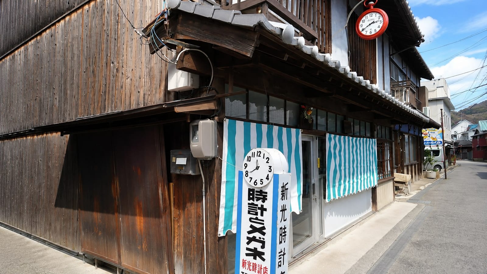 Shinko Tokeiten, a historic clock and watch repair shop in Mitarai set in a dark two-storey timber building, a large red clock mounted under the corner eaves and a painted clock sign above the striped entrance curtains.