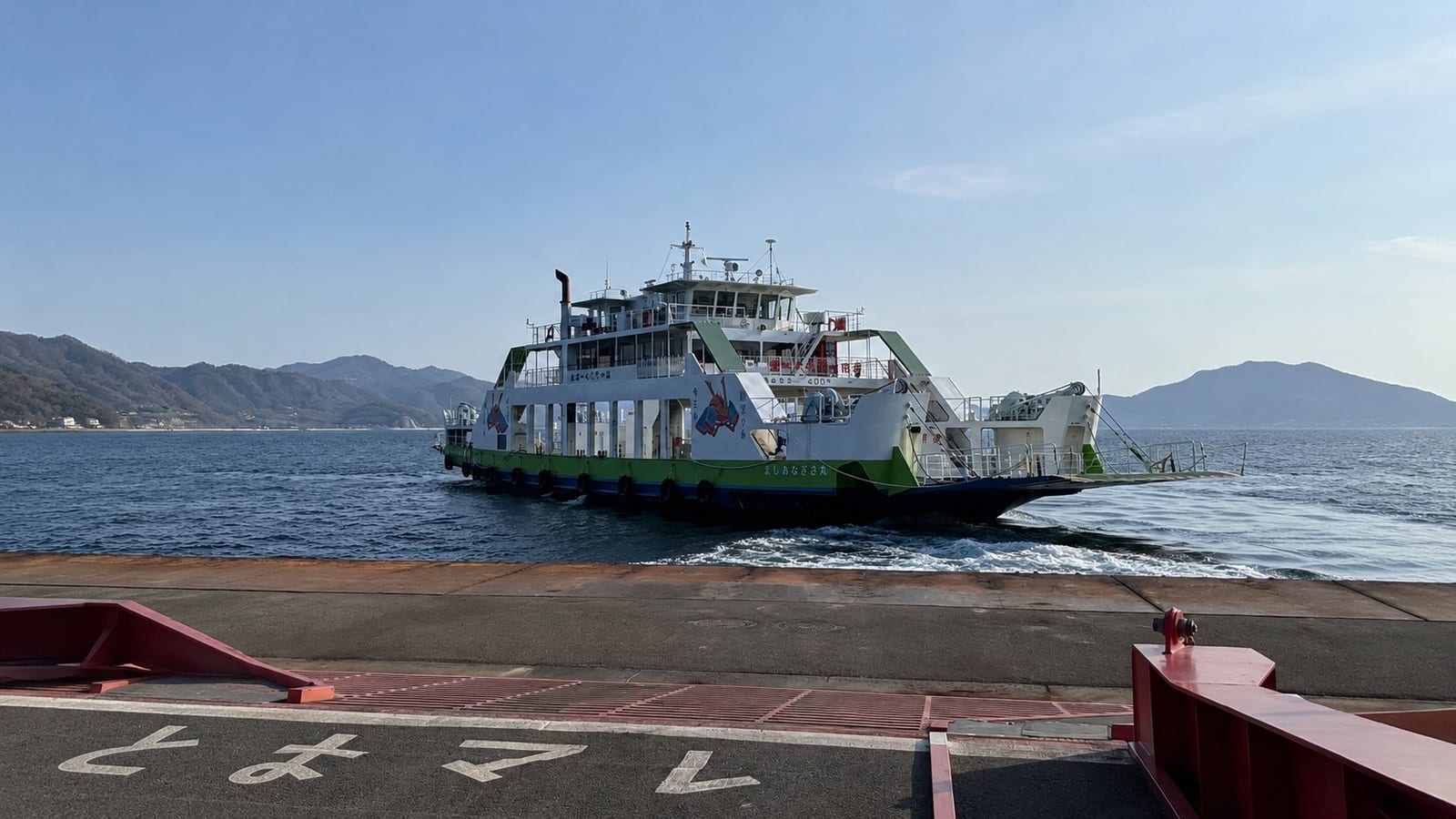 A green and white car and passenger ferry, its hull decorated with rabbit illustrations, arriving at the pier ramp on the Tadanoumi to Okunoshima route, with calm water and pine-covered island hills behind under a clear sky.