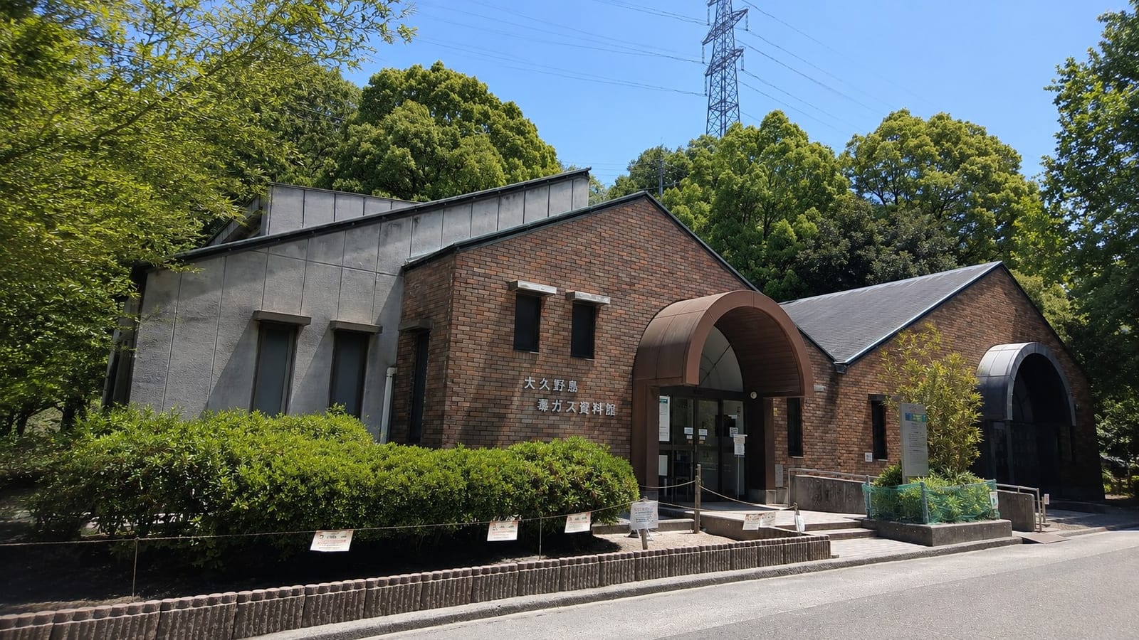 The exterior of the Okunoshima Poison Gas Museum, a modest single-storey brick building with an arched entrance and its name lettered in Japanese on the wall, set among green trees under a clear sky.
