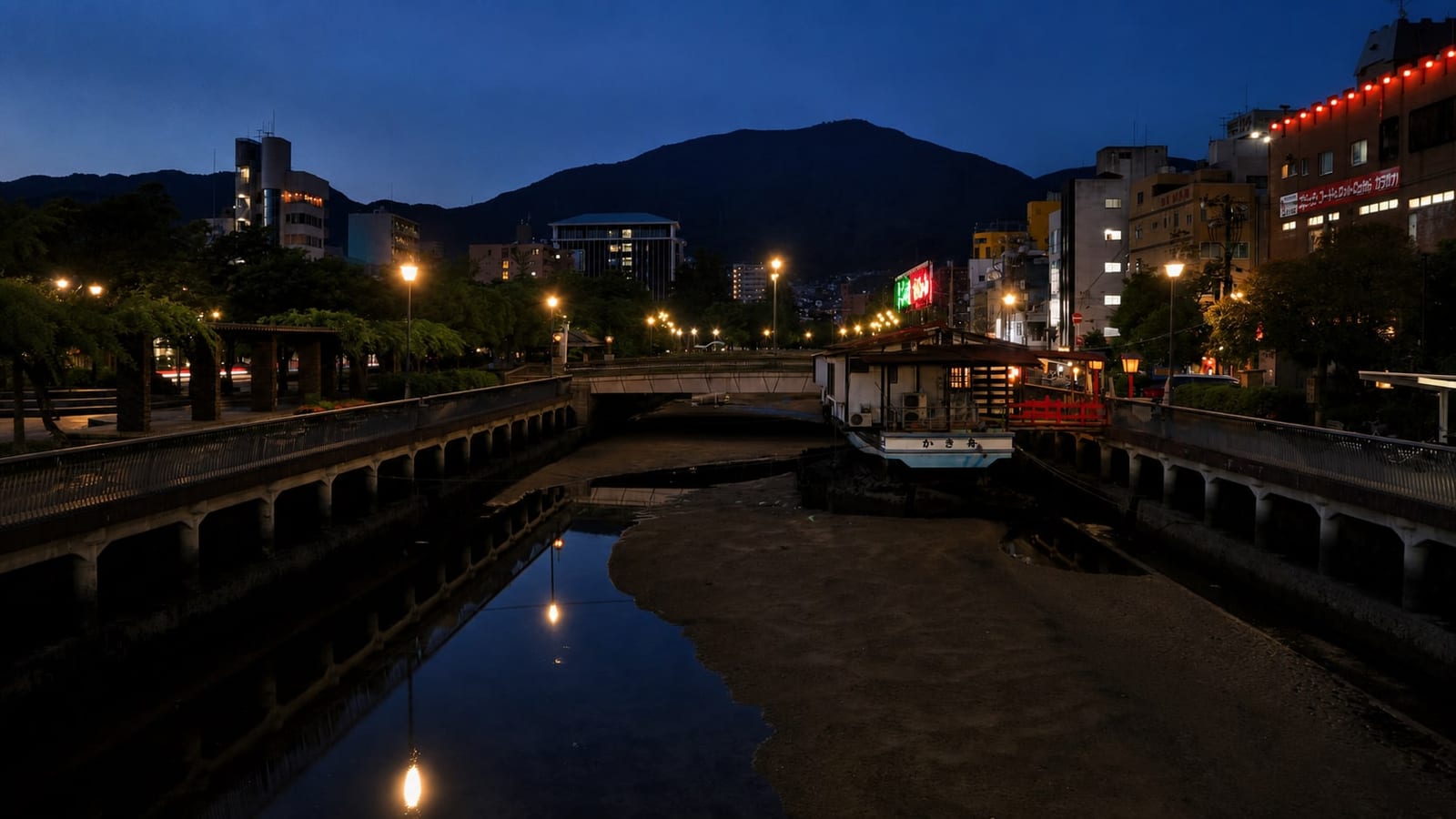 Blue hour on the Sakaigawa river in central Kure: a kakibune floating oyster restaurant moored by the bridge, street lamps reflected in the water, and Mt. Haigamine rising dark behind the city.