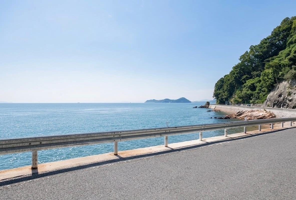 Photograph of the coastal road on the Tobishima Kaido, a narrow single-lane road along a low seawall with calm Seto Inland Sea water visible just below, small islands in the distance, overcast sky. Summer 2026.