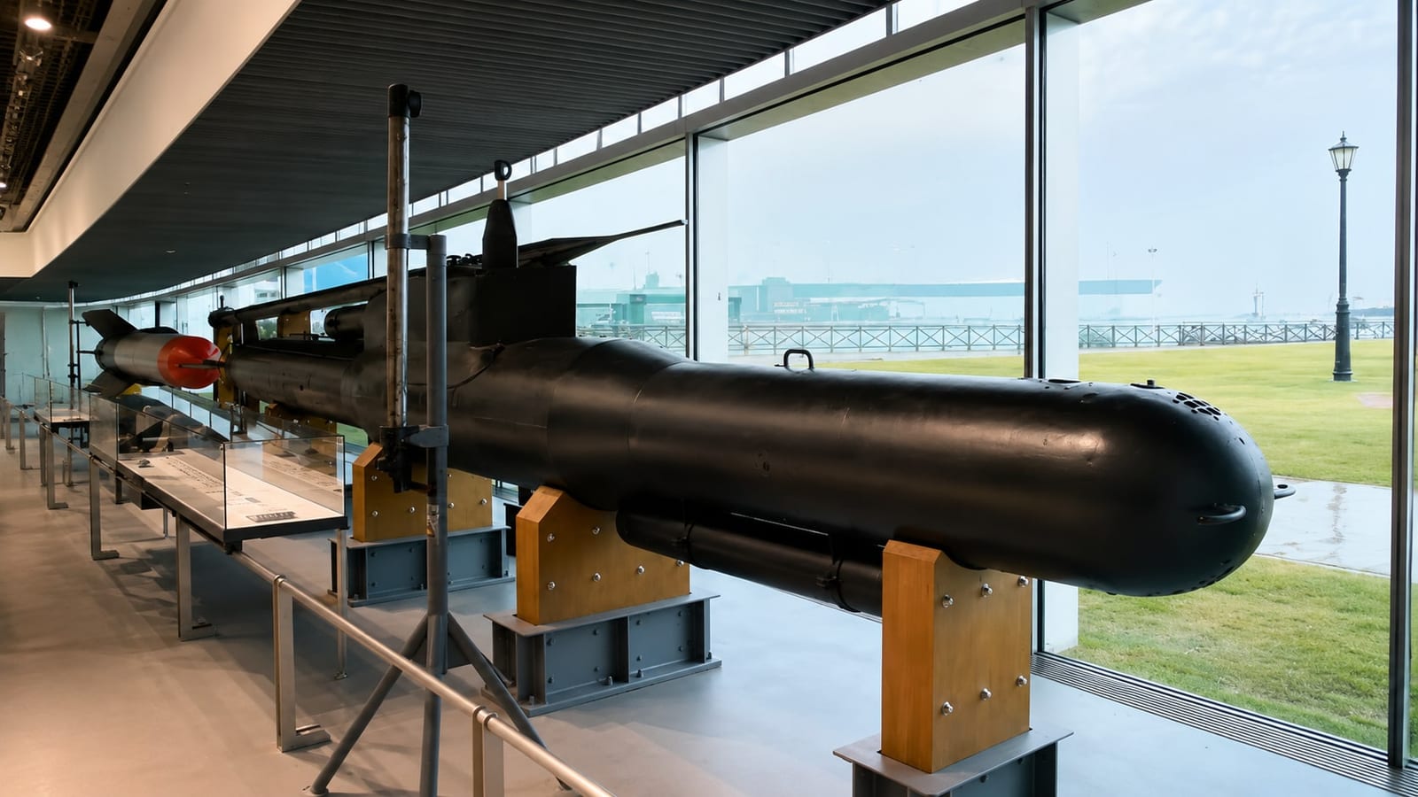 The black Type 10 Kaiten manned torpedo resting on steel and timber cradles along the glass wall of the museum's large exhibition room, with the harbour-side lawn visible outside.