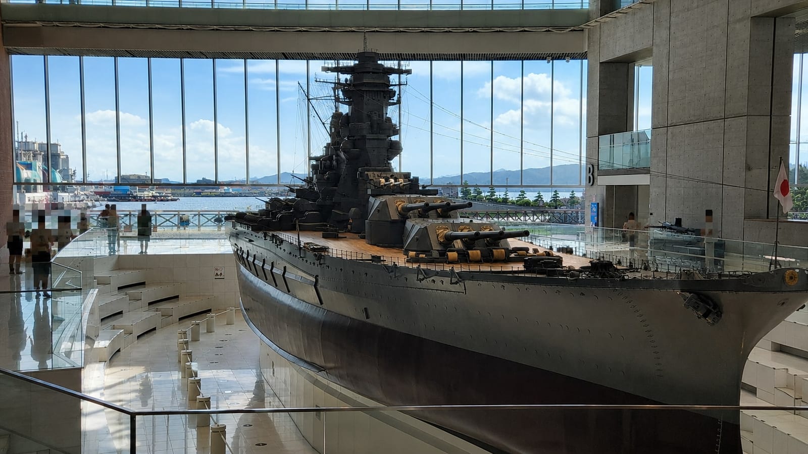 The 1:10 scale model of battleship Yamato seen from the walkway on the bow side, its main turrets and superstructure framed against the atrium's glass wall with Kure harbour behind.
