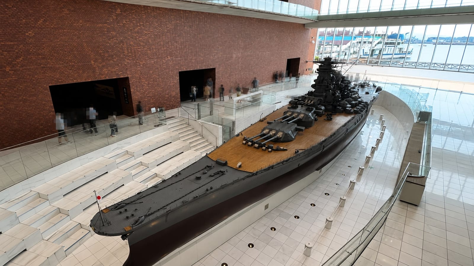 Looking down on the full 26.3-meter length of the 1:10 Yamato model from the museum's upper level, with the ferry pier visible through the glass wall behind the stern.