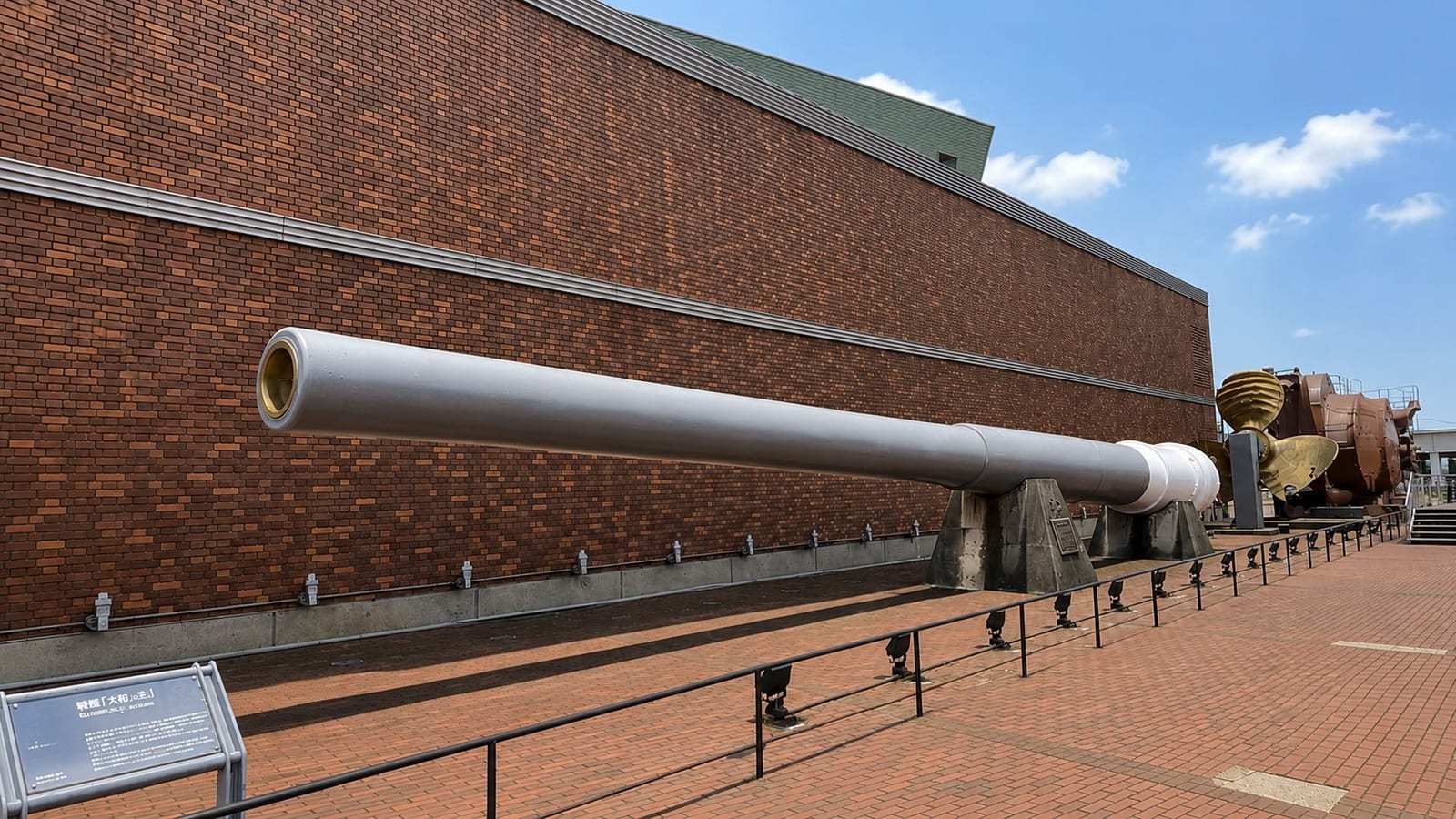 The salvaged main gun barrel of the battleship Mutsu displayed outdoors along the brick wall of the Yamato Museum, with her bronze propeller and rudder visible behind it, under a clear summer sky.