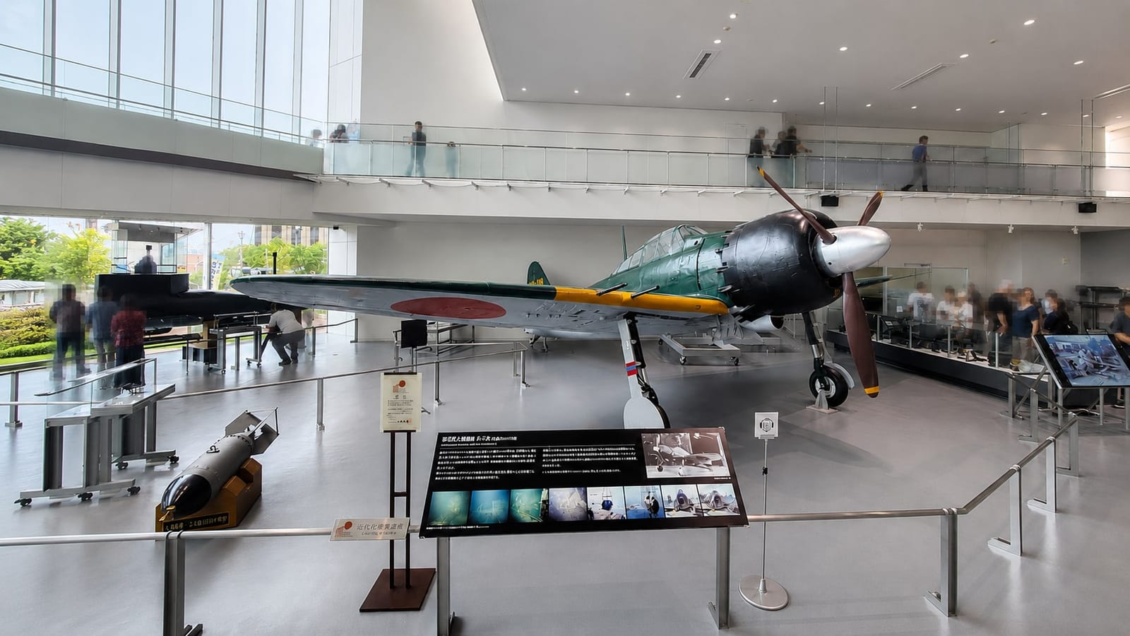 The restored Mitsubishi A6M Zero fighter in the museum's large exhibition room, seen from the front with its explanation panel in the foreground and visitors on the mezzanine above.